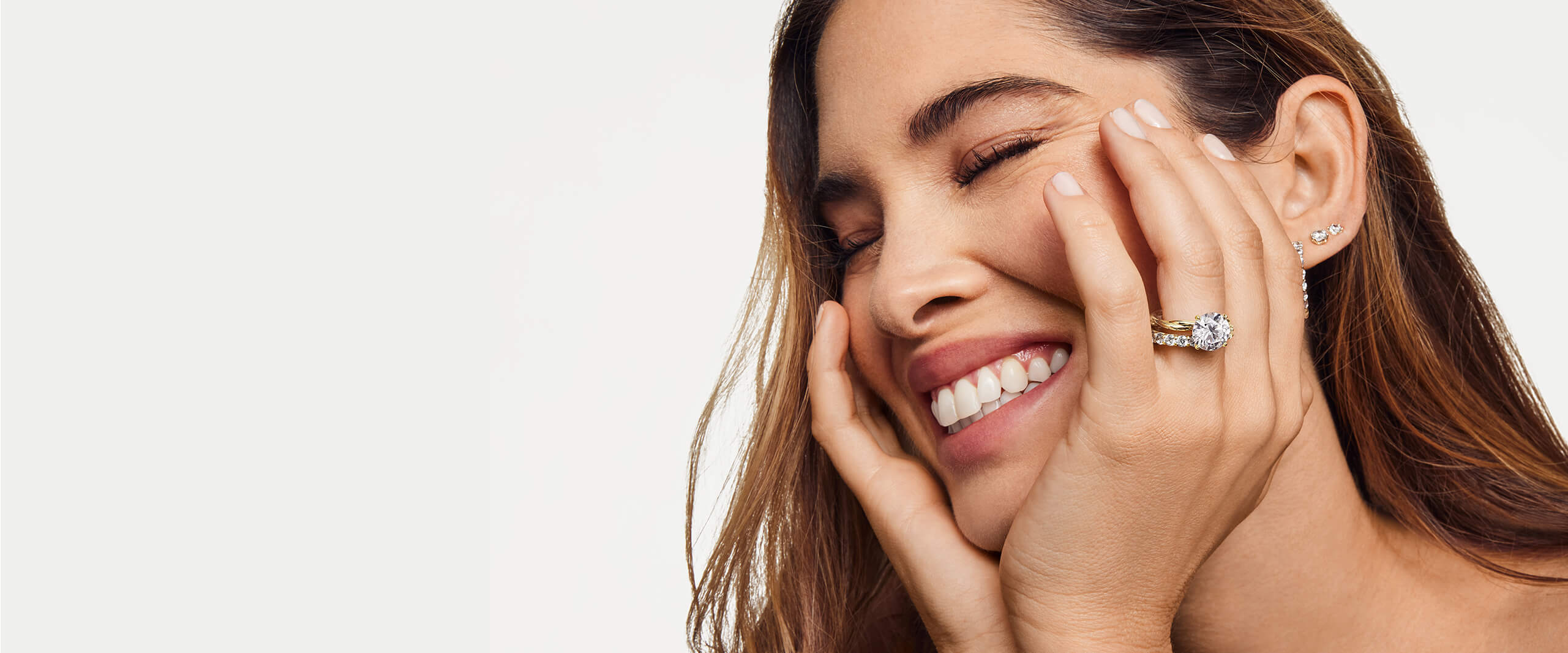 A happy woman gently cups her face with her hands, showing off her diamond accessories and large diamond engagement ring.