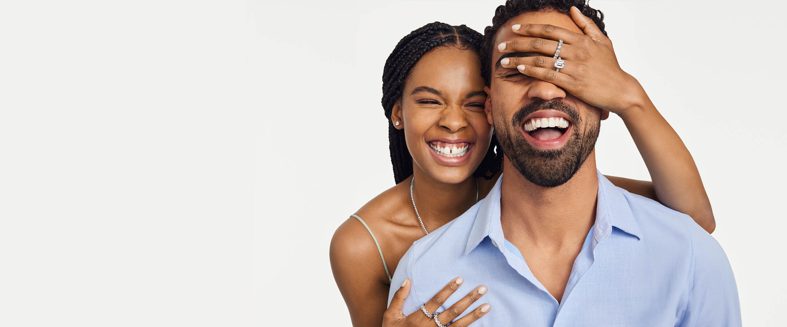 A happy couple laughs together as the woman covers her partner’s eyes while wearing an assortment of diamond jewelry and a large diamond engagement ring.